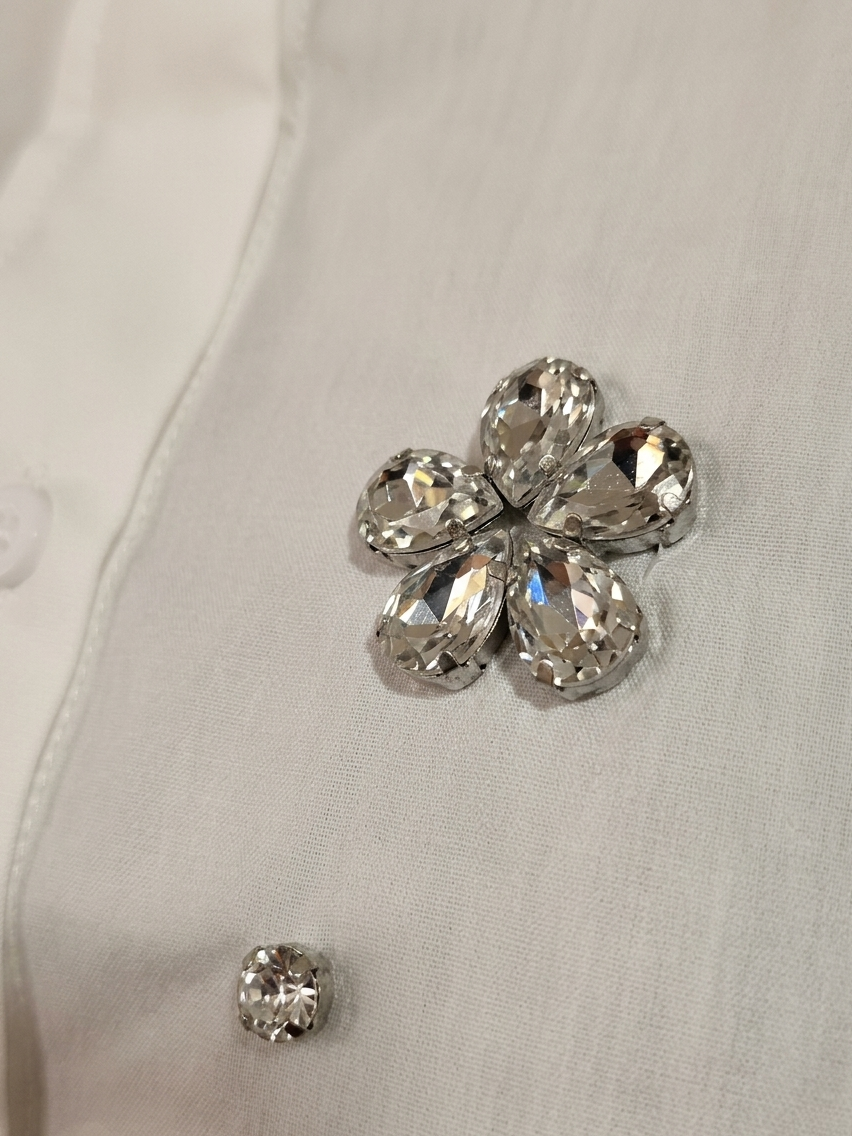 ​Front and back view of a woman wearing a white short-sleeve button-down shirt featuring crystal floral rhinestone embellishments, paired with black denim shorts. The image includes close-up shots of the 3D jewel flower appliqué and the crisp cotton fabric texture.