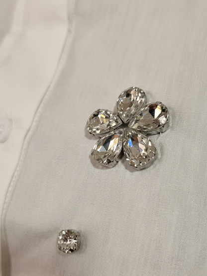 ​Front and back view of a woman wearing a white short-sleeve button-down shirt featuring crystal floral rhinestone embellishments, paired with black denim shorts. The image includes close-up shots of the 3D jewel flower appliqué and the crisp cotton fabric texture.